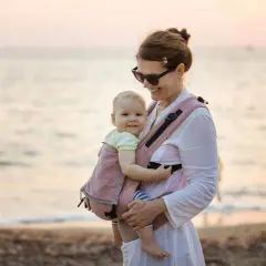 Szczęśliwa mama z dzieckiem w nosidełku na plaży. Idealne rozwiązanie dla rodziców, które mogłoby znaleźć się w rankingu nosidełek dla niemowląt.