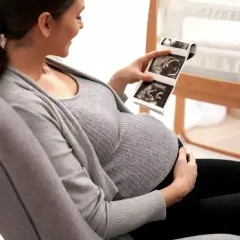 Uśmiechnięta ciężarna kobieta ogląda zdjęcia z domowego USG ciąży. W tle widać łóżeczko dziecięce.