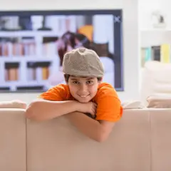 Uśmiechnięty chłopiec w czapce i pomarańczowej koszulce opiera się o kanapę. W tle telewizor i półki z książkami. Dziecko samo w domu, wiek ok. 10 lat.