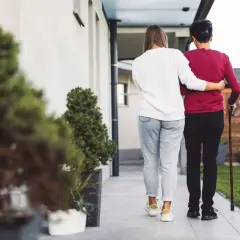 Wizyta pielęgniarki środowiskowej w domu. Młoda kobieta wspiera starszą panią z laską, idąc chodnikiem obok domu.