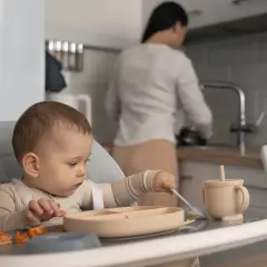 Małe dziecko siedzi w krzesełku, próbując samodzielnie jeść. Na talerzyku ma pomarańczowe kawałki owoców i łyżeczkę. To jego pierwsza kaczka dla niemowlaka.