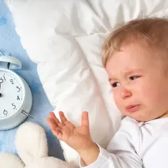 Płacz dziecka obok budzika sugeruje, dlaczego dziecko nie chce spać. Maluch leży na niebieskim kocyku z gwiazdkami, obok pluszowych zabawek.