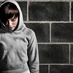 Smutna dziewczyna w szarej bluzie z kapturem stoi przy ceglanej ścianie. Może to być scena z poprawczaka dla dzieci, gdzie szuka nadziei.