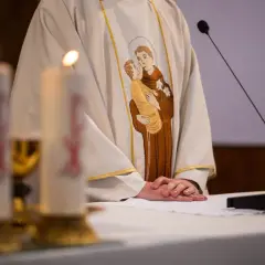 Ksiądz w szacie z haftem przedstawiającym św. Antoniego z Dzieciątkiem Jezus, przy ołtarzu ze świecami.