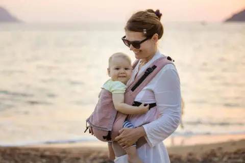 Szczęśliwa mama z dzieckiem w nosidełku na plaży. Idealne rozwiązanie dla rodziców, które mogłoby znaleźć się w rankingu nosidełek dla niemowląt.