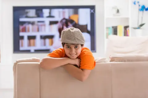 Uśmiechnięty chłopiec w czapce i pomarańczowej koszulce opiera się o kanapę. W tle telewizor i półki z książkami. Dziecko samo w domu, wiek ok. 10 lat.