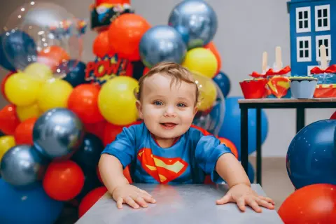Uśmiechnięty chłopiec w stroju Supermana, otoczony balonami. To dziecko ma w sobie mnóstwo radości i odwagi, co są jego mocne strony, przykłady widać w jego spojrzeniu.