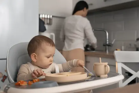 Małe dziecko siedzi w krzesełku, próbując samodzielnie jeść. Na talerzyku ma pomarańczowe kawałki owoców i łyżeczkę. To jego pierwsza kaczka dla niemowlaka.
