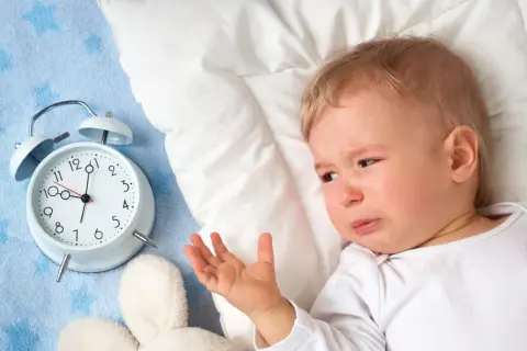 Płacz dziecka obok budzika sugeruje, dlaczego dziecko nie chce spać. Maluch leży na niebieskim kocyku z gwiazdkami, obok pluszowych zabawek.