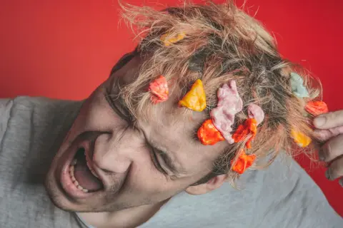 Zdezorientowany mężczyzna z gumą w blond włosach. Jak wyciągnąć gumę z włosów? To pytanie zadaje sobie, próbując ją usunąć.