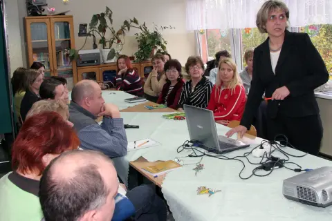 Nauczyciele zebrani na radzie pedagogicznej, dyskutują przy stole z laptopem i projektorem.