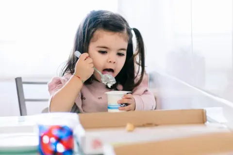 Mała dziewczynka z kitkami zajada łyżeczką pyszne danonki dla dzieci.