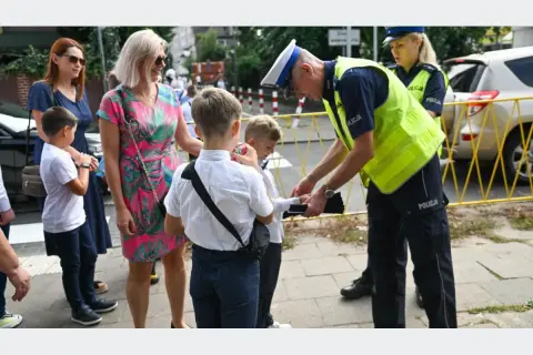 Policjant pomaga dziecku przejść przez ulicę, przypominając, od ilu lat dziecko może samo chodzić do szkoły.