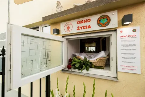 Okno życia, bezpieczne miejsce dla noworodków, jest otwarte. W koszyku czeka otulone kocykiem niemowlę, a obok bukiet czerwonych róż.