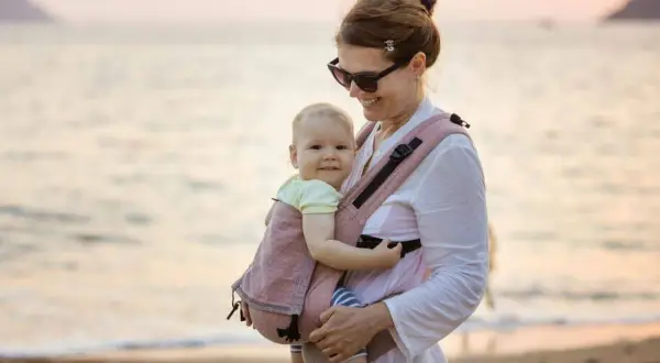 Szczęśliwa mama z dzieckiem w nosidełku na plaży. Idealne rozwiązanie dla rodziców, które mogłoby znaleźć się w rankingu nosidełek dla niemowląt.
