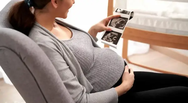 Uśmiechnięta ciężarna kobieta ogląda zdjęcia z domowego USG ciąży. W tle widać łóżeczko dziecięce.