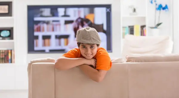 Uśmiechnięty chłopiec w czapce i pomarańczowej koszulce opiera się o kanapę. W tle telewizor i półki z książkami. Dziecko samo w domu, wiek ok. 10 lat.