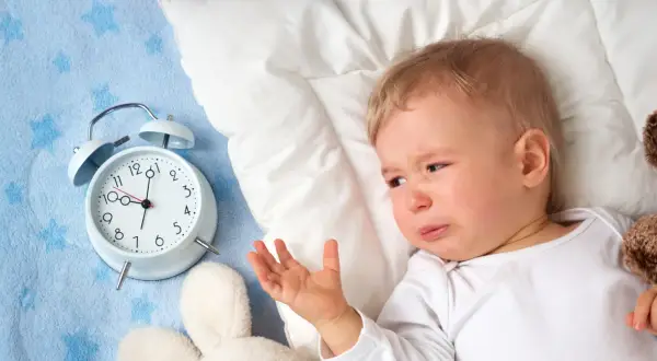Płacz dziecka obok budzika sugeruje, dlaczego dziecko nie chce spać. Maluch leży na niebieskim kocyku z gwiazdkami, obok pluszowych zabawek.
