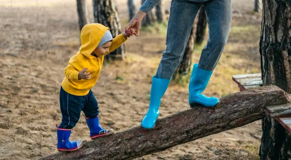 Dziecko w żółtej bluzie i mama w niebieskich kaloszach idą po kłodzie. Czy po szczepieniu można iść z dzieckiem na spacer? Tak, jeśli czuje się dobrze!