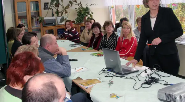 Nauczyciele zebrani na radzie pedagogicznej, dyskutują przy stole z laptopem i projektorem.