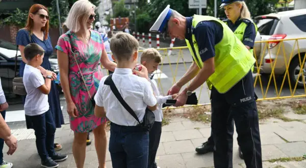 Policjant pomaga dziecku przejść przez ulicę, przypominając, od ilu lat dziecko może samo chodzić do szkoły.