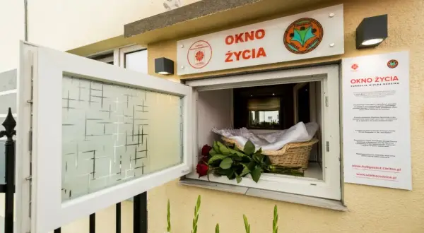 Okno życia, bezpieczne miejsce dla noworodków, jest otwarte. W koszyku czeka otulone kocykiem niemowlę, a obok bukiet czerwonych róż.