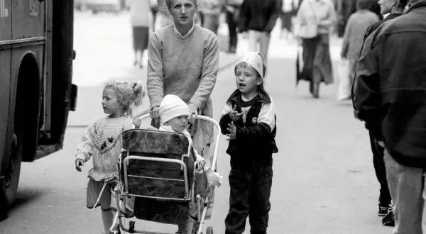 Kobieta pcha wózki dziecięce z lat 90. na ulicy, obok niej idzie starsze dziecko.