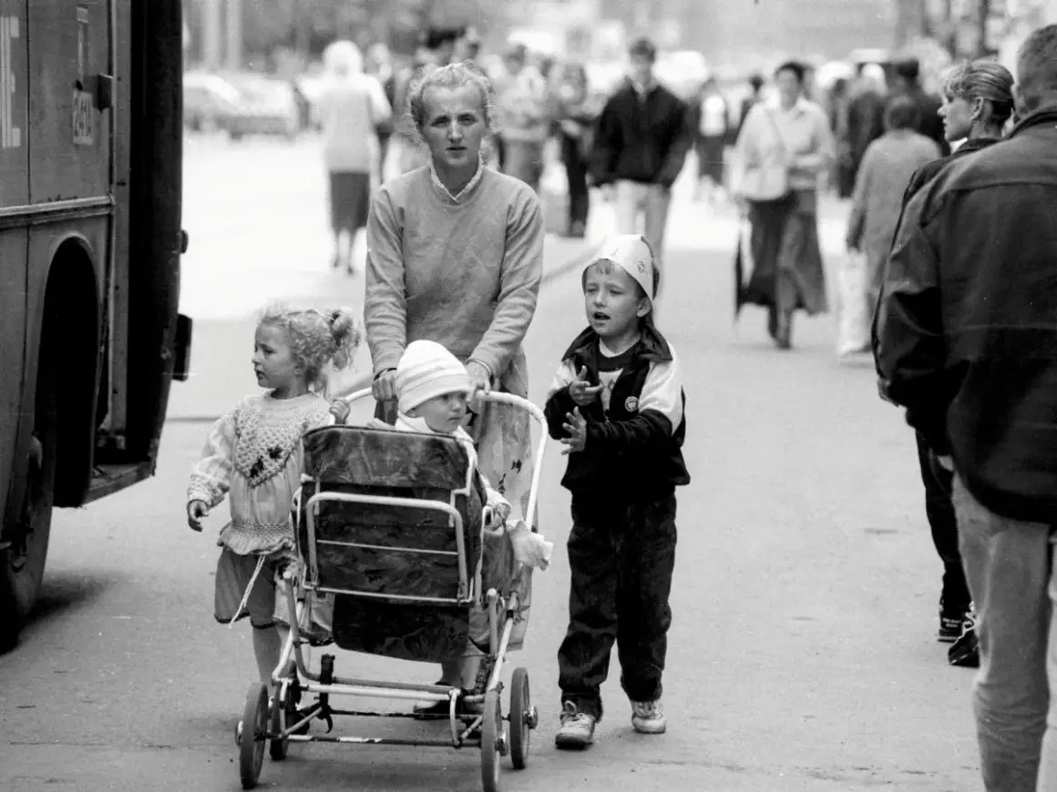Kobieta pcha wózki dziecięce z lat 90. na ulicy, obok niej idzie starsze dziecko.