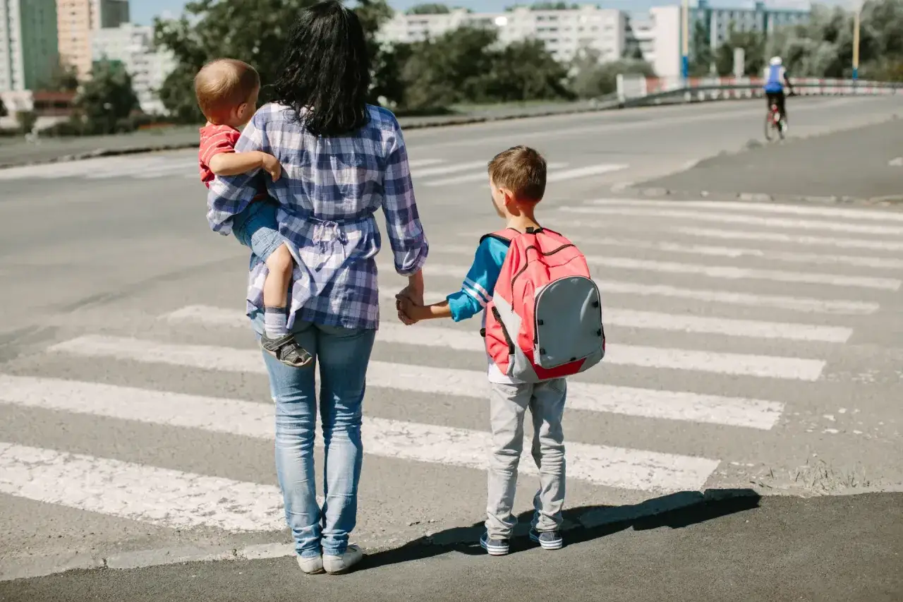 Matka z dwójką dzieci na pasach. Starsze dziecko z plecakiem, młodsze na rękach. Zastanawiamy się, od ilu lat dziecko może samo chodzić do szkoły.