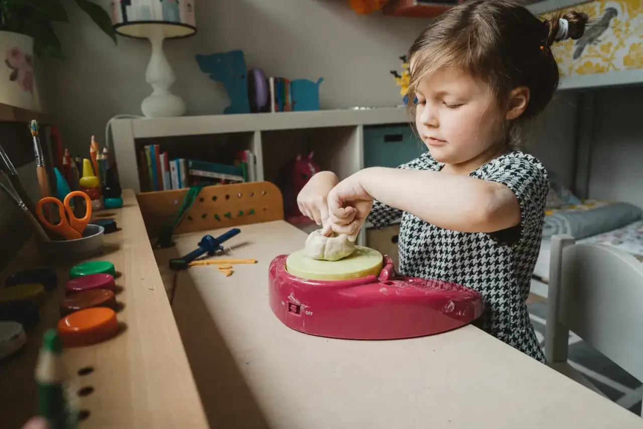 Mała dziewczynka z zapałem lepi z gliny na stoliku, gdzie leżą kolorowe kredki i nożyczki. Opis pod zdjęciem dziecka.