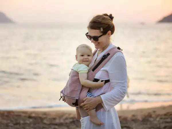 Szczęśliwa mama z dzieckiem w nosidełku na plaży. Idealne rozwiązanie dla rodziców, które mogłoby znaleźć się w rankingu nosidełek dla niemowląt.